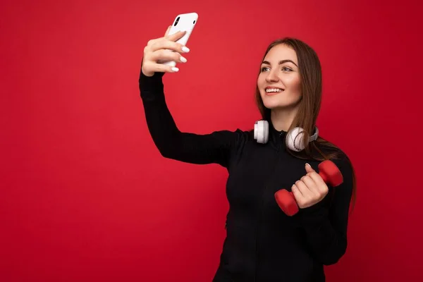 Photo of beautiful positive young brunette woman wearing black sport clothes white headphones nad holding red dumbbell standing isolated over red background using mobile phone taking selfie photo — Stock Photo, Image