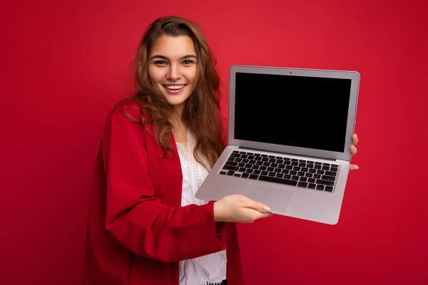 Portret de aproape de frumos zâmbind fericit tânără brunetă care deține laptop-ul computerului cu ecran de monitor gol purtând cardigan roșu și bluză albă uitându-se la camera izolată pe fundal roșu — Fotografie, imagine de stoc