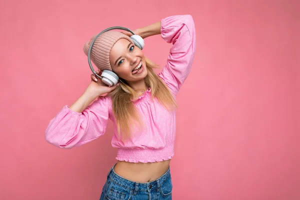 Foto de sexy atractiva mujer rubia sonriente positiva con blusa rosa y sombrero rosa aislado sobre la pared de fondo rosa con auriculares bluetooth inalámbricos blancos escuchando música que tiene — Foto de Stock