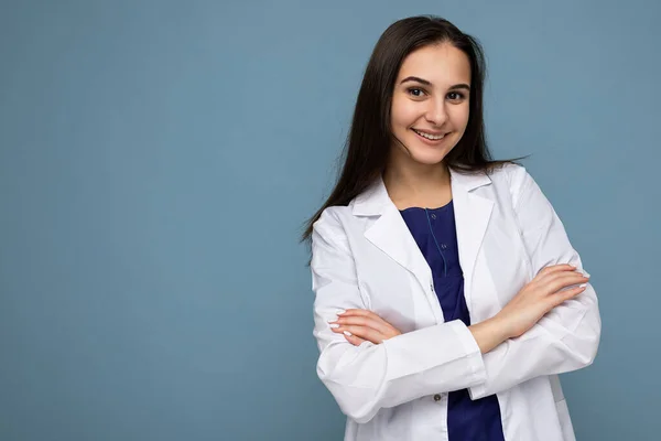 Foto retrato de jovem bonita bonita positiva sorrindo morena mulher com emoções sinceras vestindo casaco médico branco isolado sobre fundo azul com espaço de cópia e segurando os braços cruzados — Fotografia de Stock