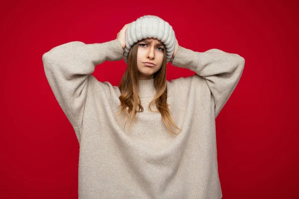 Foto de hermosa triste disgustada joven rubia oscura insatisfecha de pie aislada sobre la pared de fondo rojo con suéter beige cálido y sombrero beige de invierno mirando a la cámara — Foto de Stock