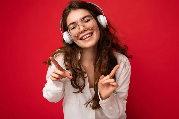 Foto de primer plano de atractiva joven morena sonriente positiva con camisa blanca y gafas ópticas aisladas sobre fondo rojo con auriculares bluetooth inalámbricos blancos escuchando música y — Foto de Stock