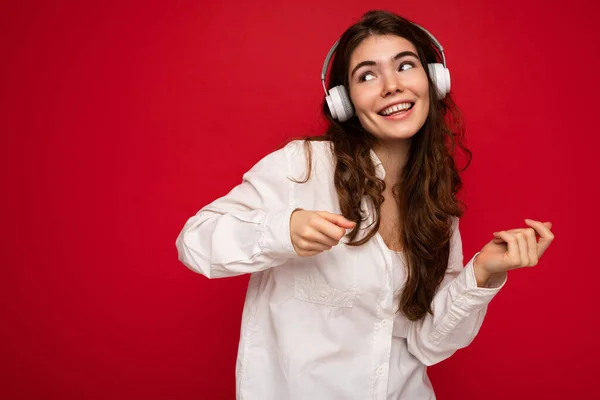 Atraktivní šťastný úsměv mladá brunetka žena osoba nosí bílou košili a optické brýle izolované přes červené pozadí na sobě bílé bezdrátové bluetooth sluchátka poslech hudby a tance — Stock fotografie