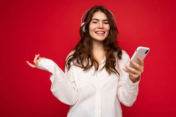 Привлекательная улыбающаяся молодая женщина в стильном повседневном наряде, изолированном на красочном фоне, держит в руках и показывает мобильный телефон с пустым экраном для выреза в белом блютузе — стоковое фото