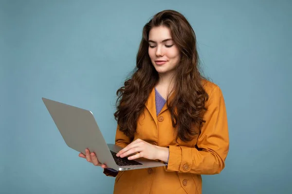 Muito morena encaracolado jovem segurando netbook computador olhando para baixo vestindo casaco amarelo digitando no teclado isolado no fundo azul — Fotografia de Stock