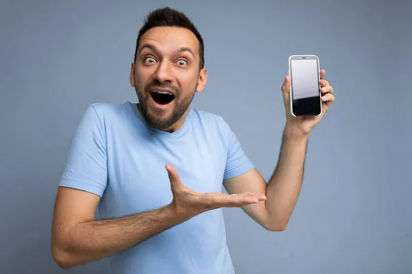 Erstaunlich gut aussehender junger unrasierter, brünetter Mann, der alltägliche blaue T-Shirts isoliert über blauem Hintergrund hält und Handy mit leerem Display zeigt, um in die Kamera zu schauen und auf — Stockfoto