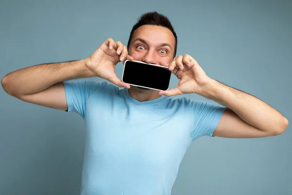 Positive handsome young unshaven brunet man with beard wearing everyday blue t-shirt isolated over blue background holding and showing mobile phone with empty display for mockup looking at camera — Stock Photo, Image