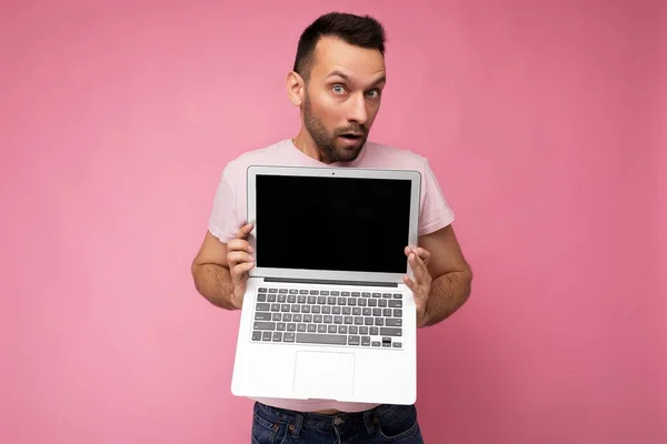 Красивий здивований і здивований чоловік тримає ноутбук, дивлячись на камеру в футболці на ізольованому рожевому фоні — стокове фото