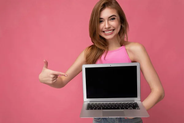 Schöne junge blonde Frau hält Computer-Laptop mit leerem Monitor-Display und zeigt auf Netbook mit rosa Crop Top, die Kamera isoliert auf rosa Hintergrund — Stockfoto