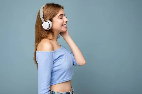 Side-profile photo of beautiful positive smiling young blonde woman wearing blue crop top isolated over blue background wall wearing white wireless bluetooth headsets listening to cool music and — Stock Photo, Image