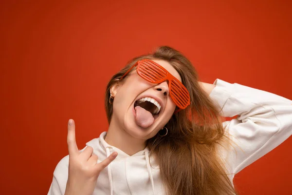 Shot of beautiful crazy overjoyed young blonde woman isolated over colourful background wearing casual white capodie and stylish sunglasses and showing rock and roll sign — Stock fotografie