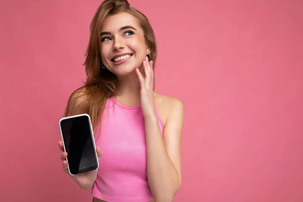 섹시하고 행복 한 젊은 금발 여성 이 분홍색 맨 위를 입고 빈 공간을 손에 쥐고 핸드폰을 보여 주며 흉내를 내며 핸드폰을 보여 주고 있습니다. — 스톡 사진