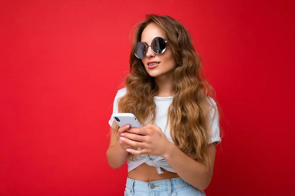 Φωτογραφία από ελκυστική θετική εμφάνιση νεαρή γυναίκα φορώντας casual κομψό ντύσιμο poising απομονωμένη στο παρασκήνιο με κενό χώρο που κατέχουν στο χέρι και τη χρήση sms κινητό τηλέφωνο ψάχνει να — Φωτογραφία Αρχείου