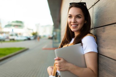 Mutlu, hoş görünümlü, çekici, büyüleyici, büyüleyici, neşeli, neşeli, düz saçlı, sokağın kenarında dizüstü bilgisayar ve beyaz tişörtlü gözlüklü bir kadın.