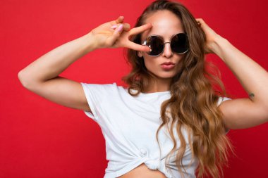 Kırmızı arka plan duvarından izole edilmiş, beyaz tişört ve şık güneş gözlüğü takmış, kameraya bakıp barış jesti yapan büyüleyici, genç, esmer, kıvırcık saçlı kadının yakın plan fotoğrafı.