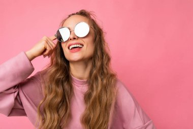 Güzel, mutlu, gülümseyen, esmer, kıvırcık saçlı bir kadının pembe arka plan duvarında izole edilmiş, günlük pembe spor giysileri ve şık güneş gözlükleri takmış bir fotoğrafı.