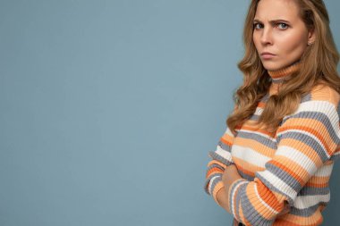 Genç, güzel, üzgün, mutsuz, samimi duyguları olan, mavi arka planda izole edilmiş, sıradan çizgili kazak giyen, mutsuz, sarışın bir kadının portresi.