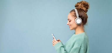 Beyaz bluetooth kulaklıklar ve beyaz kulaklıklar takarak renkli arka plan duvarından izole edilmiş şık giyinen güzel, neşeli, güler yüzlü, genç bir kadının panoramik yan plan fotoğrafı.