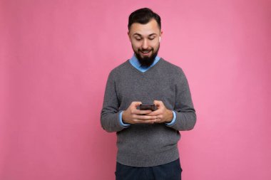 Fotoğrafta güzel görünümlü, sakallı, sakalsız, gri kazaklı, mavi gömlekli, pembe arka plan duvarına izole edilmiş, elinde boş bir alan olan ve cep telefonu kullanan yakışıklı bir adam görülüyor.