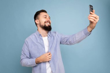 Yakışıklı, şık kıyafetler giyen arka plandaki duvarda izole bir şekilde duran akıllı telefon taşıyan cep telefonu ekranına bakarak selfie çeken genç bir adam.