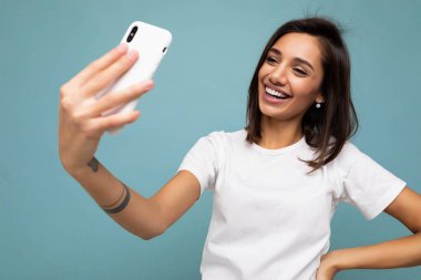 Çekici, genç, gülümseyen, mutlu bir kadın cep telefonu tutuyor ve kullanıyor. Selfie çekiyor. Duvar arkasından izole edilmiş şık kıyafetler giyiyor.