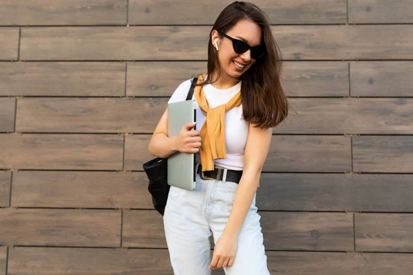 Fascinating beautiful smiling charming young brunet girl looking down holding computer laptop and black sunglasses in white t-shirt and light blue jeans in the street near brown wall — Stock Photo, Image