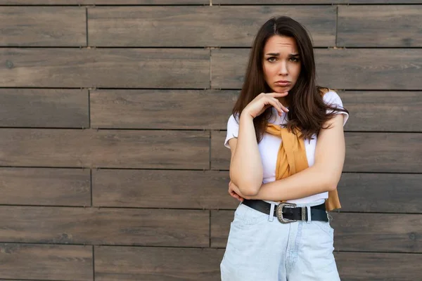 Portrét nedůtklivé smutné rozrušené uražené mladé brunetky žena na sobě ležérní bílé tričko a džíny se žlutým svetrem balancující v blízkosti hnědé zdi na ulici — Stock fotografie
