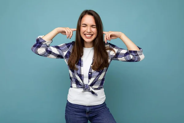 Je ne veux pas l'entendre. Jeune femme brune émotionnelle positive et souriante portant une chemise à carreaux isolée sur fond bleu avec espace de copie et oreilles couvrantes — Photo