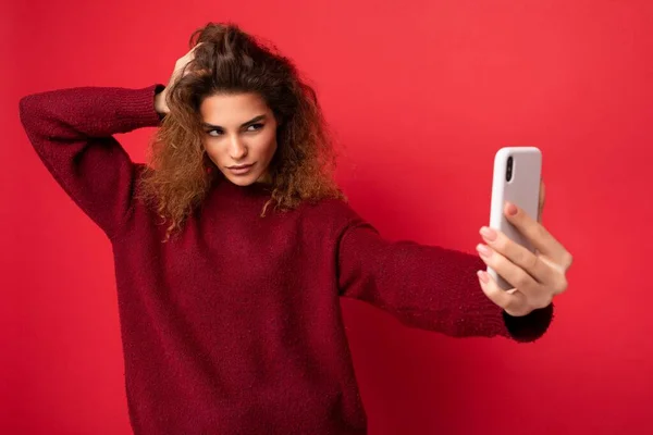 Sexy Attractive young blogger woman with curly hair wearing dark red sweater isolated on red background wall holding and using smart phone looking at telephone screen and taking selfie — Stock Photo, Image
