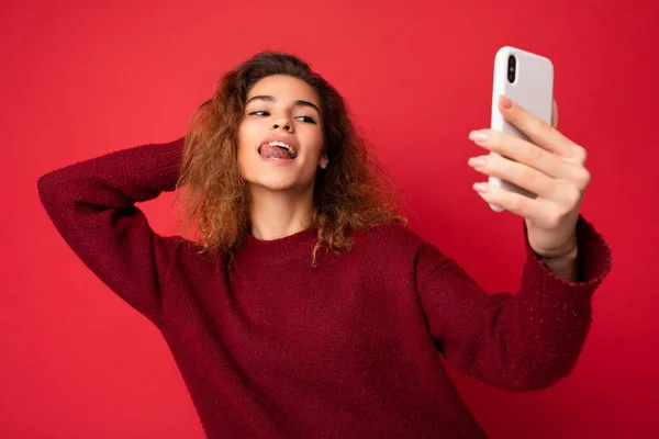 Mulher jovem sexual bonita com cabelo encaracolado vestindo camisola vermelha escura isolada na parede de fundo vermelho segurando e usando telefone inteligente olhando para a tela do telefone e tomando selfie e mostrando a língua — Fotografia de Stock