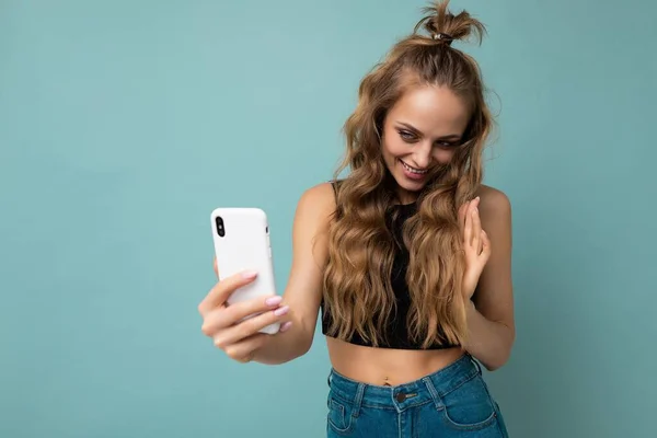 Atractiva encantadora joven sonriente mujer feliz sosteniendo y usando el teléfono móvil tomando selfie usando ropa elegante aislado sobre fondo de pared — Foto de Stock
