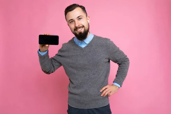 Feliz sonriente guapo feliz fresco joven morena sin afeitar hombre con barba usando elegante suéter gris y camisa azul de pie aislado sobre fondo rosa de la pared sosteniendo el teléfono inteligente y mostrando el teléfono — Foto de Stock