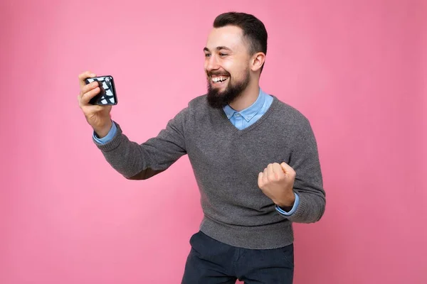 拍摄快乐英俊的年轻黑发男子，未刮胡子，身穿日常灰色毛衣和蓝色衬衫，被隔离在背景墙上，手持智能手机通过手机观看视频 — 图库照片