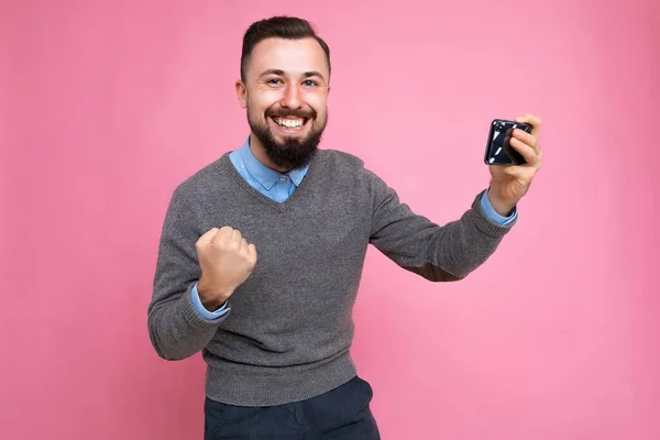 매일 회색 스웨터와 파란 셔츠를 입은 행복하고 잘생긴 젊은 갈색 머리의 남자가 스마트폰을 통해 비디오를 보고 있는 배경 벽에 고립되어 있는 사진. — 스톡 사진