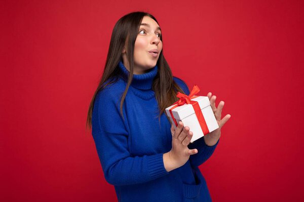 Shot of attractive happy smiling sexy young brunette woman isolated over red background wall wearing blue casual sweater holding white gift box and looking to the side