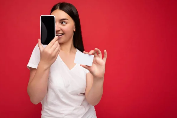 Крупный план довольно счастливая молодая брюнетка женщина в повседневной белой футболке изолированы на красном фоне с пустым местом, держа в руке мобильный телефон и показывая смартфон с пустым экраном для — стоковое фото