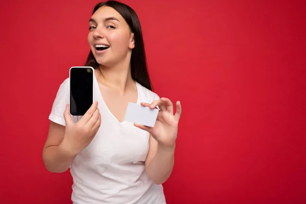 Sexy attractive happy young brunette woman wearing casual white t-shirt isolated over red background with empty space holding in hand mobile phone and showing smartphone with empty screen for mockup — Stock Photo, Image