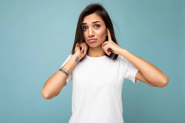 Retrato de una joven guapa morena con emociones sinceras usando una camiseta blanca casual para maqueta aislada sobre fondo azul con espacio para copiar y cubrir las orejas con los dedos — Foto de Stock