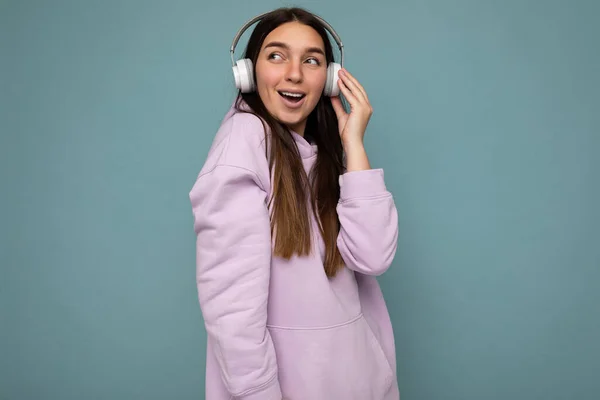 Hermosa feliz sonriente joven morena persona femenina con sudadera con capucha púrpura luz aislada sobre la pared de fondo azul con auriculares bluetooth blancos escuchando música fresca y mirando a un lado — Foto de Stock