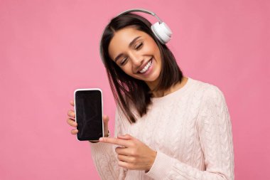 Pembe süveter giyen, güzel, genç, esmer bir kadın. Pembe arka plan duvarından izole edilmiş, beyaz bluetooth kulaklık takıyor, iyi müzik dinliyor ve cep telefonunu boş gösteriyor.