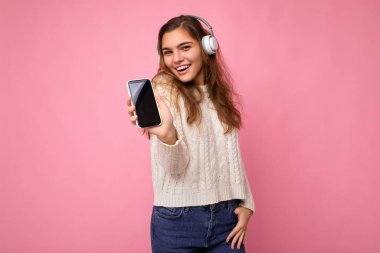 Güzel, mutlu, güler yüzlü, şık kıyafetler giyen, arka plandaki duvarda izole edilmiş, cep telefonlu beyaz kulaklık takan, model için boş bir ekran gösteren genç bir kadın.