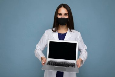 İleri profil fotoğrafı, beyaz önlüklü ve siyah maskeli genç esmer kadın elinde dizüstü bilgisayarla mavi duvarda izole edilmiş kameraya bakıyor.