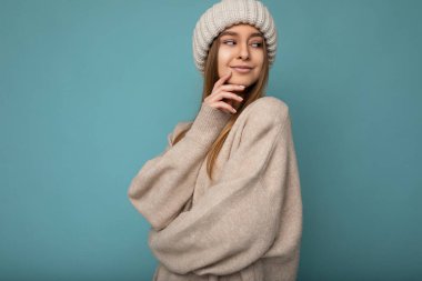 Çekici, mutlu, esmer, sarışın bir kadının, mavi arka plan duvarında izole edilmiş, bej kazak ve kış bej şapkalı bir şekilde duruşu.