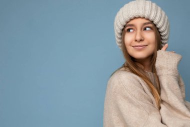 Güzel, gülümseyen, sarışın bir kadının, mavi arka plan duvarında izole bir şekilde durduğu, bej kazak ve kış bej şapkası giydiği bir fotoğraf.