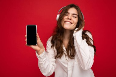 Çekici, pozitif, kıvırcık saçlı, beyaz gömlek giyen, kırmızı arka plan duvarında izole edilmiş, cep telefonunu boş bir şekilde gösteren, beyaz kulaklık takan, mavi kulaklık takan bir kadın.