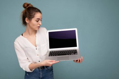 Netbook bilgisayarı tutan güzel, gülümseyen genç bir kadının yan profil portresi mavi arka planda beyaz gömlek giyen monitöre bakıyor.