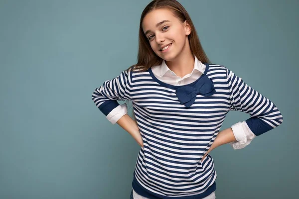 Foto de hermosa niña morena sonriente positiva con manga larga a rayas de pie aislado sobre la pared de fondo azul y mirando a la cámara — Foto de Stock