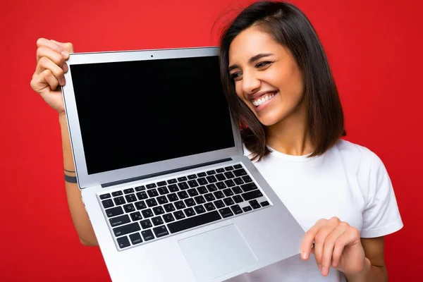 Fotografie de zâmbet amuzant Frumos fericit copleșit femeie tânără cu părul scurt brunet care deține laptop-ul computerului în căutarea laterală purtând tricou alb și blugi izolați pe fundalul peretelui roșu — Fotografie, imagine de stoc
