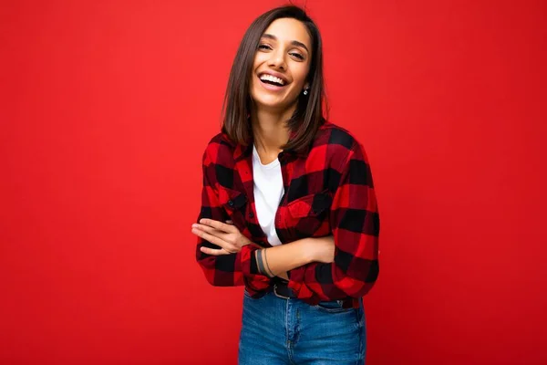 Портрет позитивной веселой улыбчивой молодой брюнетки в повседневной белой футболке, стильная красная чековая рубашка, выделенная на красном фоне с копировальным пространством — стоковое фото