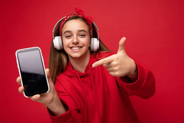 Foto de una linda morena sonriente positiva con capucha roja aislada sobre fondo rojo sosteniendo y mostrando el teléfono inteligente con pantalla vacía para recortar usando auriculares inalámbricos blancos escuchando — Foto de Stock